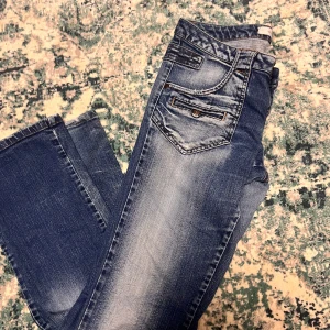 Blå jeans med slitningar - Säljer ett par snygga blå jeans med coola slitningar och detaljerade fickor. De har en klassisk femficksdesign och är perfekta för en avslappnad stil. Passar både till vardags och fest!