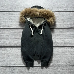 Svart hoodie med fuskpäls zip up y2k swag - Snygg svart hoodie med dragkedja och huva kantad med fuskpäls. Perfekt för kyliga dagar med sin mjuka insida och ribbade muddar. Den har isolerad insida, passar skitbra och är knappt använd. Bredden är 51 och längden 60.