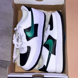 Nike Air Force med gröna detaljer - Snygga Nike Air Force sneakers i vitt med svarta och gröna detaljer. Skorna har en klassisk design med snörning och en bekväm passform. Perfekta för en stilren look.