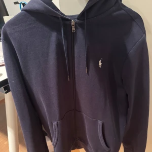 Mörkblå Ralph Lauren zip up hoodie - Säljer en mörkblå hoodie från Ralph Lauren med dragkedja och klassisk logga på bröstet. Perfekt för en avslappnad look. Den har en bekväm passform och praktiska fickor framtill. Skriv om ni har några frågor! Priset är ej hugget i sten.