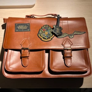 Brun axelväska med Hogwarts-motiv - Brun axelväska i skinn med Hogwarts- motiv från Primark. Väskan har två framfickor och en justerbar axelrem. Perfekt för Harry Potter-fans💕