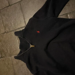 Svart stickad tröja från Polo Ralph Lauren - Snygg svart stickad tröja från Polo Ralph Lauren med dragkedja och röd broderad logga på bröstet. Perfekt för kyligare dagar med sin ribbade krage och muddar.