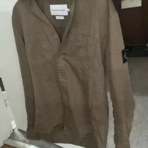 Brun skjorta från Calvin Klein Jeans - Snygg brun skjorta från Calvin Klein Jeans med lång ärm och klassisk krage. Skjortan har en bröstficka och en stilren logga på ärmen. Perfekt för en avslappnad men stilren look.