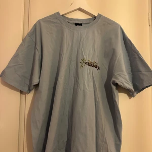 Ljusblå t-shirt från Stussy - Snygg ljusblå t-shirt från Stussy med blommigt mönster runt loggan både fram och bak. Perfekt för en avslappnad stil. T-shirten har en normal passform och korta ärmar. Strl M men oversized så passar L