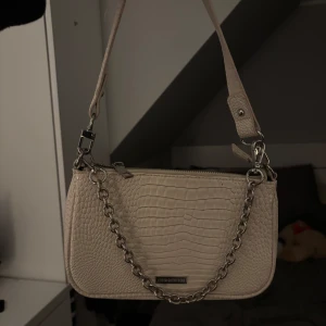 Beige axelväska från Urban Outfitters - Snygg beige axelväska från Urban Outfitters med krokodilpräglat mönster. Väskan har en kedjedetalj och dragkedja upptill. Perfekt för att ge en stilren touch till din outfit.