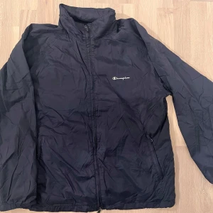 Svart vindjacka från Champion - Säljer en svart vindjacka från Champion med dragkedja och hög krage. Jackan har en enkel design med Champion-loggan på bröstet och praktiska fickor med dragkedja. Perfekt för blåsiga dagar.