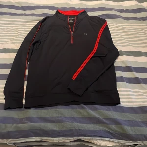 Svart tröja med dragkedja från Calvin Klein - Snygg svart tröja från Calvin Klein med röda detaljer och dragkedja. Tröjan har långa ärmar och en liten CK-logga på bröstet. Perfekt för en stilren look. Pris går alltid att förhandlas! 