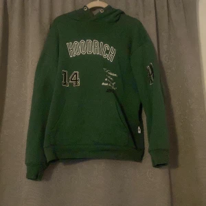 Grön hoodie från Hoodrich - Säljer en grön hoodie från Hoodrich med texten 'HOODRICH' och siffran '14' tryckt på framsidan. Den har en klassisk huva och långa ärmar, perfekt för en avslappnad stil.