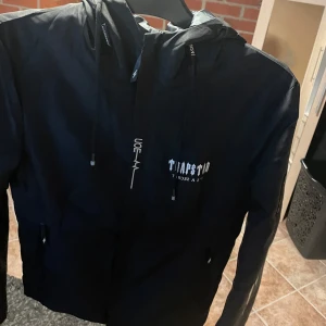 Svart vindjacka från Trapstar - Snygg svart vindjacka från Trapstar med huva och dragkedja. Jackan har tryck på både fram- och baksidan. Perfekt för blåsiga dagar.