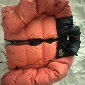 Rosa dunjacka från The North Face - Säljer en snygg rosa dunjacka från The North Face med svarta detaljer. Jackan har en dragkedja framtill och är perfekt för kyliga dagar. Den är både stilren och funktionell, vilket gör den till ett måste i garderoben.