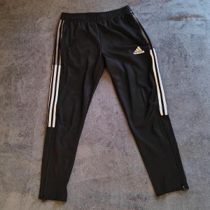 Träningsbyxor Adidas - Svarta träningsbyxor från Adidas med klassiska vita ränder längs sidorna. Köpt i Adidas butiken på Sveavägen Stockholm för 500 kr. Byxorna har en elastisk midja och dragkedjor vid bensluten för extra komfort. Perfekta för att träna och gymma i, eller för att slappna av hemma. Liten missfärgning på linjerna men syns knappt (bild 5). Hör av dig vid frågor eller funderingar!
