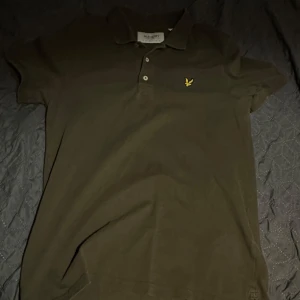 Olivgrön pikétröja från Lyle & Scott - Säljer en olivgrön pikétröja från Lyle & Scott med korta ärmar och klassisk krage. Tröjan har en broderad gul logotyp på bröstet och knappar framtill. Perfekt för en avslappnad stil.