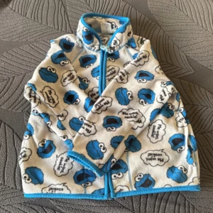 Vit och blå fleecejacka med mönster - Säljer en mysig vit fleecejacka med blått mönster av tecknade figurer. Jackan har en dragkedja framtill och är långärmad med blå kantdetaljer. Perfekt för kyliga dagar!