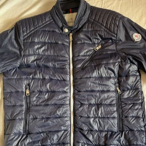 Blå pufferjacka från Moncler - Snygg blå pufferjacka från Moncler med quiltad design och dragkedja framtill. Jackan har en broderad logotyp på ärmen och praktiska fickor med dragkedjor. Det är storlek 3 men jag är 185 och den passar inte. Säljer för ett lägre pris för vet inte om den är äkta eller fejk och vid snabba affär.