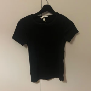 Svart ribbad kortärmad topp från H&M - Säljer en stilren svart ribbad kortärmad topp från H&M. Perfekt basplagg som passar till det mesta. Toppen har en rund halsringning och är tillverkad i ett mjukt material. Den är i mycket bra skick men används några gånger.
