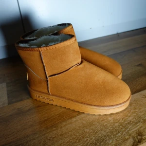 Bruna boots med foder - Mysiga bruna boots i mocka med fluffigt vitt foder. Perfekta för kalla dagar när du vill hålla fötterna varma och bekväma. De har en enkel design utan snörning, vilket gör dem lätta att ta på och av. Passar perfekt till både vardags och avslappnade tillfällen.
