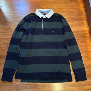 Randig pikétröja från Tommy Hilfiger - Snygg långärmad pikétröja från Tommy Hilfiger med breda ränder i mörkgrön och marinblå. Tröjan har en klassisk vit krage och knappslå framtill. Perfekt för en avslappnad stil med en touch av elegans.