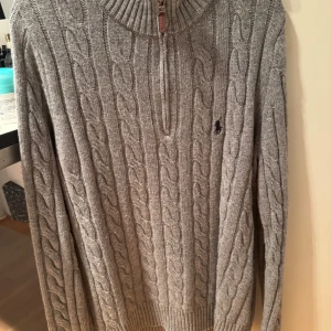 Grå kabelstickad halfzip från Polo Ralph Lauren - Säljer en stilren grå kabelstickad tröja från Polo Ralph Lauren. Tröjan har en halv dragkedja och ribbstickad krage, perfekt för kyliga dagar. Den klassiska loggan är broderad på bröstet. Inga defekter eller något sådant, pris kan diskuteras