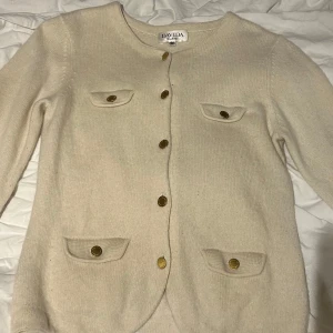 Beige kashmirkofta från Davida - Säljer en elegant beige kashmirkofta från Davida. Koftan har långa ärmar och knäpps med stiliga guldknappar. Den har fyra fickor med knappdetaljer för en klassisk look. Perfekt för en stilren och bekväm outfit.