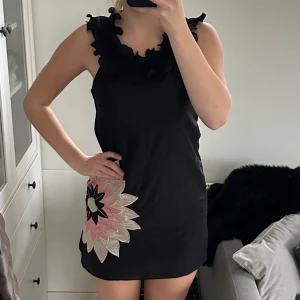 Svart klänning med volangdetaljer från Iseg - Snygg svart klänning med volangdetaljer vid axlarna och en stor blommig applikation i rosa och beige på sidan. Klänningen är ärmlös och har en rak passform som ger en stilren look. Dold dragkedja i midjan