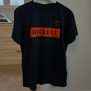 Svart t-shirt från Hackett - Snygg svart t-shirt från Hackett med orange tryck på bröstet. Perfekt för en avslappnad stil. Kortärmad och bekväm passform.