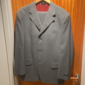 Grå kostym från Studio - Snygg grå italiensk kostym byxor och kavaj från Studio med kavaj och matchande byxor. Kavajen har klassisk design med knappar och fickor, och är fodrad med röd insida. Perfekt för formella tillfällen. Inköpt 3999kr mitt pris 949kr. Grymt skick väldigt sällan använd.