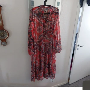 Mönstrad långklänning från Lindex - Säljer en färgglad och mönstrad långklänning från Lindex. Klänningen har långa ärmar och ett livfullt mönster i rött och andra färger. Perfekt för en bohemisk stil.