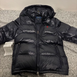 Polo Ralph Lauren jacka  - Snygg, bekväm och trendig mörkblå dunjacka från Ralph Lauren. Jackan är i perfekt skick förutom skada(baksidan höger) där materialet är ihop skrynklat. Det känns och syns inte vid användning alls. Perfekt för kyliga dagar med sin vadderade design. Kvitto medföljer. Hör av dig vid funderingar😁