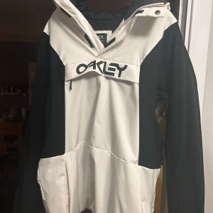 Vit och svart jacka från Oakley - Säljer en stilren vit och svart jacka från Oakley. Jackan har en stor logotyp på framsidan och en praktisk dragkedja. Perfekt för kyligare dagar med sin långa ärmar och funktionella design.