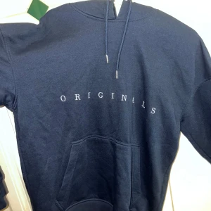 Svart hoodie från Jack & Jones - Snygg svart hoodie från Jack & Jones med texten 'ORIGINALS' på framsidan. Den har en klassisk känguruficka och justerbar huva med dragsko. Perfekt för en avslappnad stil.