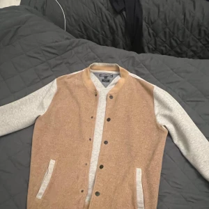 Beige och grå bomberjacka TOMMY HILFIGER 9/10 skick  - Snygg bomberjacka från Tommy hilfiger i beige och grått. Jackan har en klassisk design med knappar framtill och kontrasterande ärmar. Perfekt för en avslappnad stil.