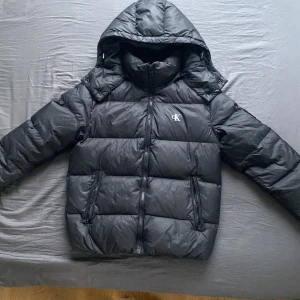 Svart dunjacka från Calvin Klein Jeans - Säljer nu min snygga svarta dunjacka från Calvin Klein med huva och dragkedja. Perfekt för kyliga dagar med sin pufferdesign som håller dig varm. Jackan har en stilren look med CK-logga på bröstet och på vänster arm. Endast använd ett få tal gånger. Bara höra av er vid frågor eller ytligare bilder. Priset går att diskutera vid snabb affär. 