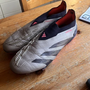 Adidas Predator fotbollsskor - Säljer ett par Adidas Predator fotbollsskor i grått med svarta och röda detaljer. Skorna har en strumpliknande passform utan snörning och en sula med röda dobbar för optimalt grepp på planen. Använda några gånger men är i mycket bra skick, ingen påse eller box tillkommer! Hojta till vid frågar🫣😁