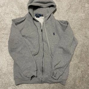 Grå hoodie från Ralph Lauren - Säljer en klassisk grå hoodie från Ralph Lauren med dragkedja och huva. Den har en broderad logga på bröstet och ribbade muddar vid ärmslut och nederkant. Perfekt för en avslappnad stil. Helt ny har ej använt, priset kan diskuteras 