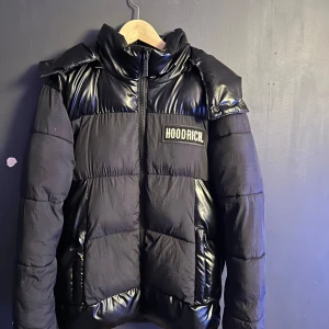 Svart pufferjacka från Hoodrich - Säljer en snygg svart pufferjacka från Hoodrich i storlek XS men passar S inte alls XS är själv 172 och den känns lite lång på mig men inget man tänker på så mycket. Jackan har en glansig finish med dragkedja och en stor huva. Perfekt för kalla dagar med sin varma vaddering. Stort Hoodrich-tryck på bröstet och ryggen ger en cool look. Passar perfekt för vintern och hösten!