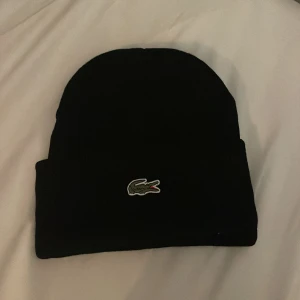 Svart mössa från Lacoste - Snygg svart mössa från Lacoste med det ikoniska krokodilemblem broderat framtill. Perfekt för att hålla värmen under kyliga dagar.  Helt ny 