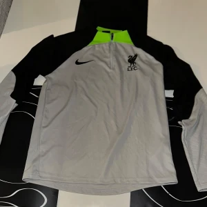 Grå och svart träningströja från Nike - Självklart! Här är en beskrivning du kan använda:  Liverpool FC Nike Träningströja Säljer en snygg och bekväm Liverpool FC Nike träningströja i storlek (S 	•	Färg: Grå med svarta ärmar och neongröna detaljer 	•	Halv zip-modell för justerbar ventilation 	•	Officiell Nike-produkt med LFC-logga och Nike Swoosh 	•	Perfekt för träning eller vardagsbruk   Tröjan är onvänd köpte den för 800kr