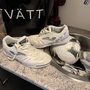Vita fotbollsskor från Joma - Säljer ett par vita fotbollsskor från Joma med snörning och perforerade detaljer för bättre ventilation. Skorna har en sportig design med grå och svarta accenter. Riktiga futsal skor som är i mycket bra skick, använda några gånger men har absolut inga fel! De är ganska stora i storleken! Så om du har vanligtvis 43,44 i storlek passar dessa bäst 