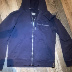  Calvin Klein tröja  - Säljer en snygg mörkblå hoodie från Calvin Klein Jeans med dragkedja och broderad logga på bröstet. Perfekt för en avslappnad stil. Den har en praktisk huva och fickor framtill.