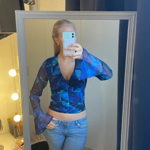 Blå mönstrad festtopp - Säljer en snygg blå långärmad festtopp med ett fint mönster. Toppen har långa ärmar och är tillverkad i ett genomskinligt material vid ärmarna. Perfekt för att sticka ut i mängden! Passar mig som bär storlek S. Använd 1 gång.