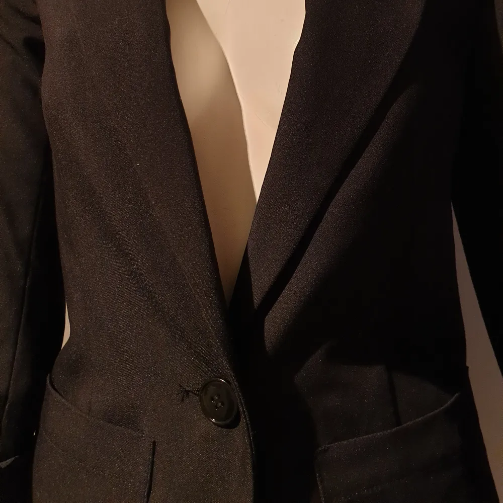 Elegant svart kavaj med klassisk design och knappar framtill. Kavajen har långa ärmar och fickor på sidorna, vilket ger en stilren look. Perfekt för en formell eller professionell stil.. Puvut & Bleiserit.