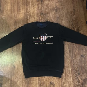 Svart sweatshirt från GANT - Säljer en stilren svart sweatshirt från GANT med broderad logga och texten 'American Sportswear' på framsidan. Tröjan har långa ärmar och en rund halsringning. Perfekt för en avslappnad stil.