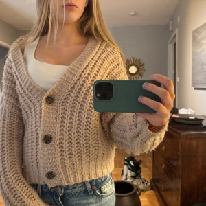 Beige stickad kofta - Säljer en mysig beige stickad kofta med stora knappar framtill. Koftan har en djup v-ringning och långa ärmar, perfekt för kyligare dagar. Den är både stilren och bekväm, idealisk för lager-på-lager-stil.