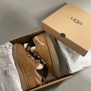 Bruna sneakers från UGG - Säljer ett par snygga bruna sneakers från UGG med snörning och mjukt foder. Skorna har en robust sula och är perfekta för kyligare dagar. De är tillverkade i mocka och har en stilren design med UGG-loggan på sidan.