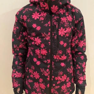 Svart och rosa blommig skidjacka  - Fickor både utanpå, intill och på vänster bröst. Rymlig luva så hjälmen får plats under.