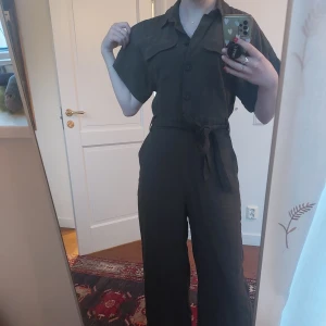 Olivgrön jumpsuit från H&M - Säljer en stilren olivgrön jumpsuit från H&M med korta ärmar och knytband i midjan. Har fållat om benen för att förlänga dem lite, och den har lite slitage längst ner vid fållen (se bild) dock inget som syns vid användning eller påverkar hållbarheten. Storlek 34 men passar mig som är 36.