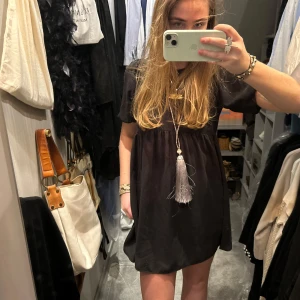 Svart kortklänning - Säljer en svart kortklänning med en unik tofsdetalj framtill. Klänningen har korta ärmar och en lös passform, perfekt för en avslappnad stil. Den är enkel men stilren och kan lätt matchas med olika accessoarer.