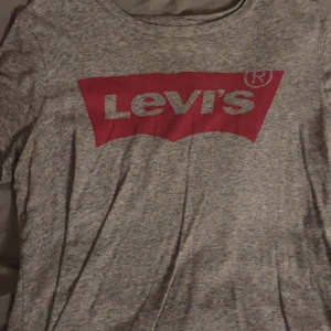 Grå t-shirt från Levi's - Säljer en klassisk grå t-shirt från Levi's med det ikoniska röda logotyptrycket på bröstet. Perfekt för en avslappnad stil. T-shirten är kortärmad och tillverkad i mjukt material.