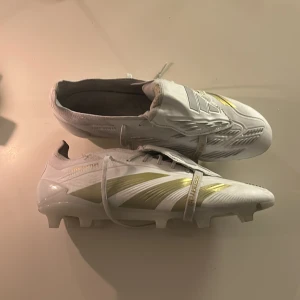 Vita Adidas Predator fotbollsskor - Säljer ett par vita Adidas Predator fotbollsskor med guldiga detaljer. Skorna har en modern design med snörning och är perfekta för fotbollsplanen. De kommer med en praktisk vit påse med Predator-logga. Använt 2 gånger