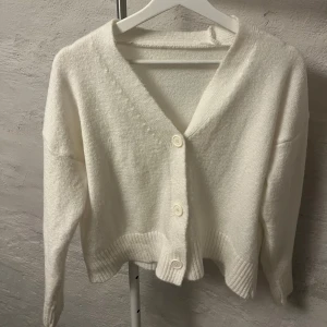 Vit kofta med knappar - Säljer en mysig vit kofta med knappar framtill. Den har en v-ringad design och långa ärmar. Perfekt för kyligare dagar.